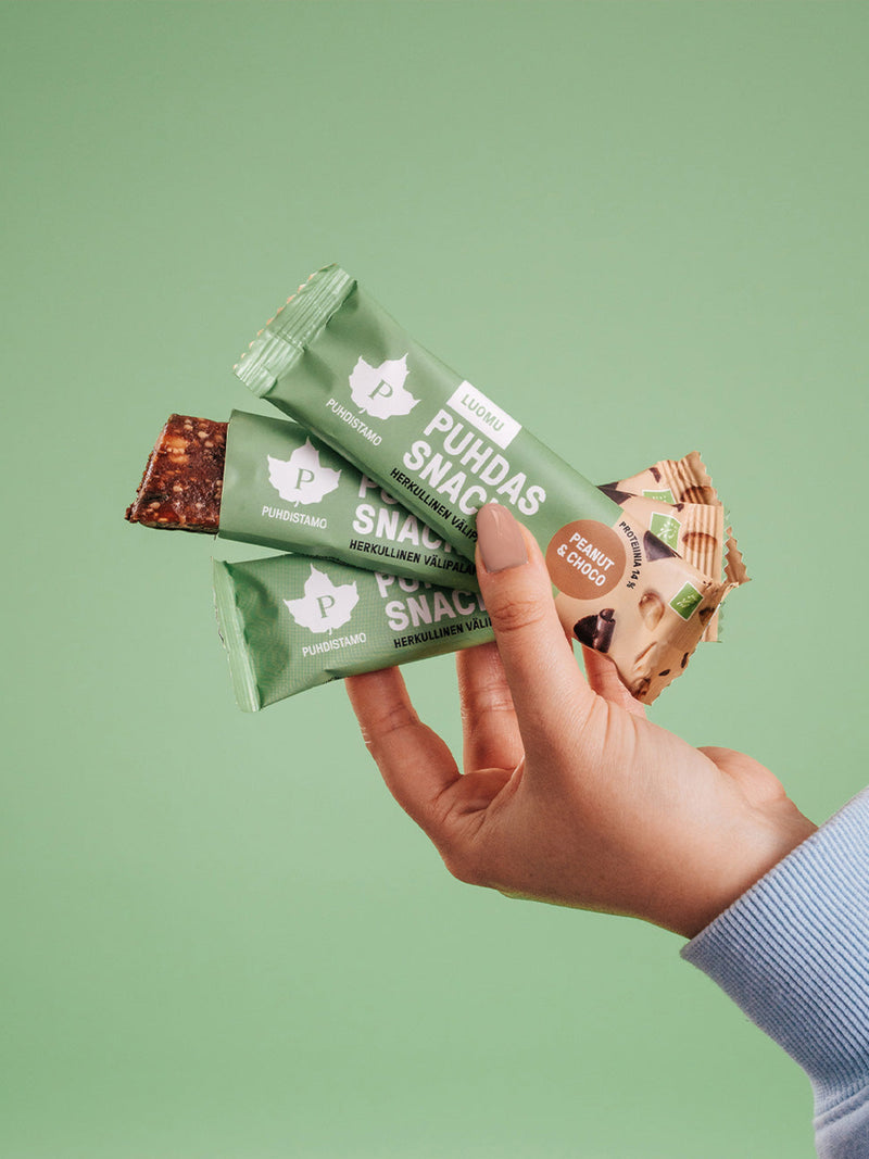 Nainen pitää kädessään kolmea Puhdistamon Puhdas Snack Välipalapatukkaa vihreää taustaa vasten