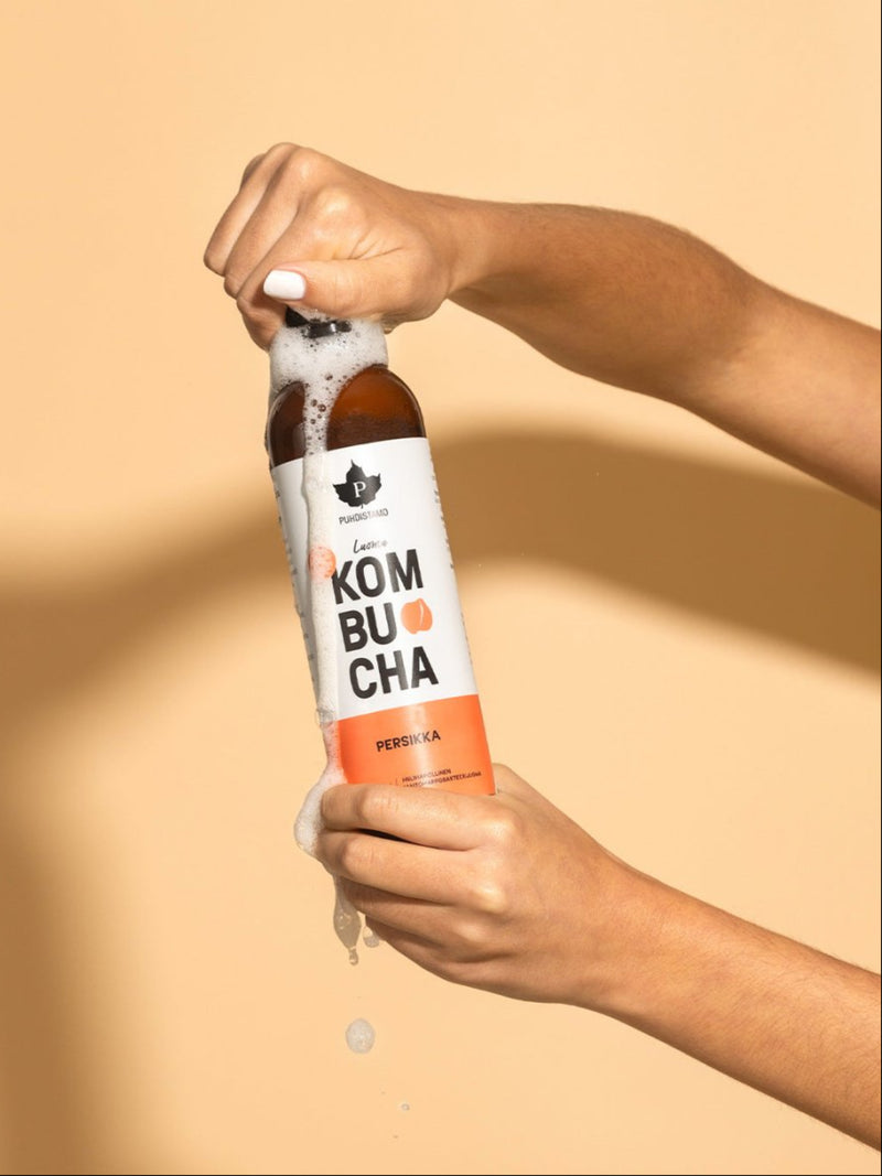 Nainen pitää kädessään kuohuavaa Puhdistamon Luomu Kombucha persikka pulloa